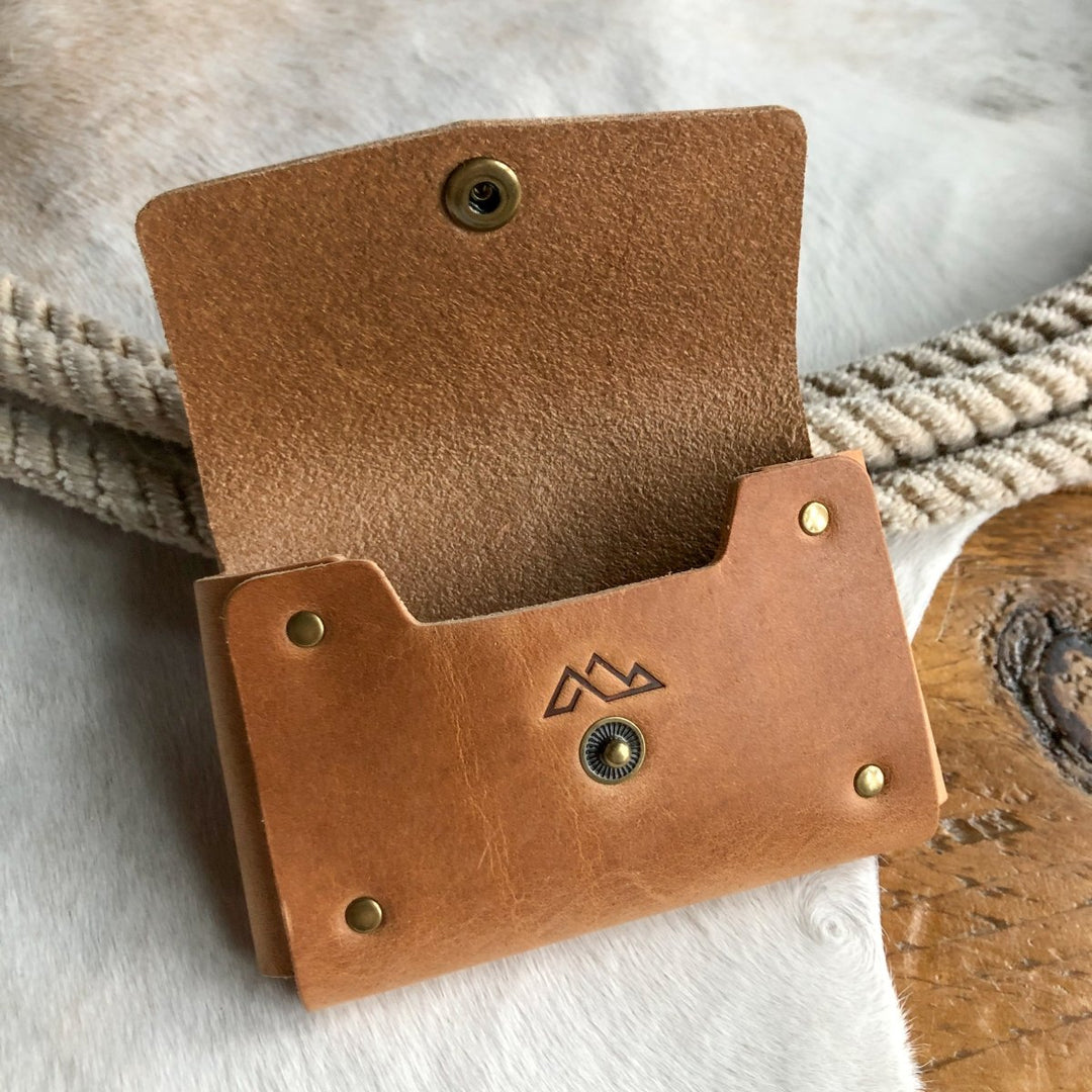 The Sorrell Card Holder by Savannah Sevens western life{&}style, made from tan Horween leather with brass rivets and a stamped mountain design, is open on a white textured surface near braided rope and wood.