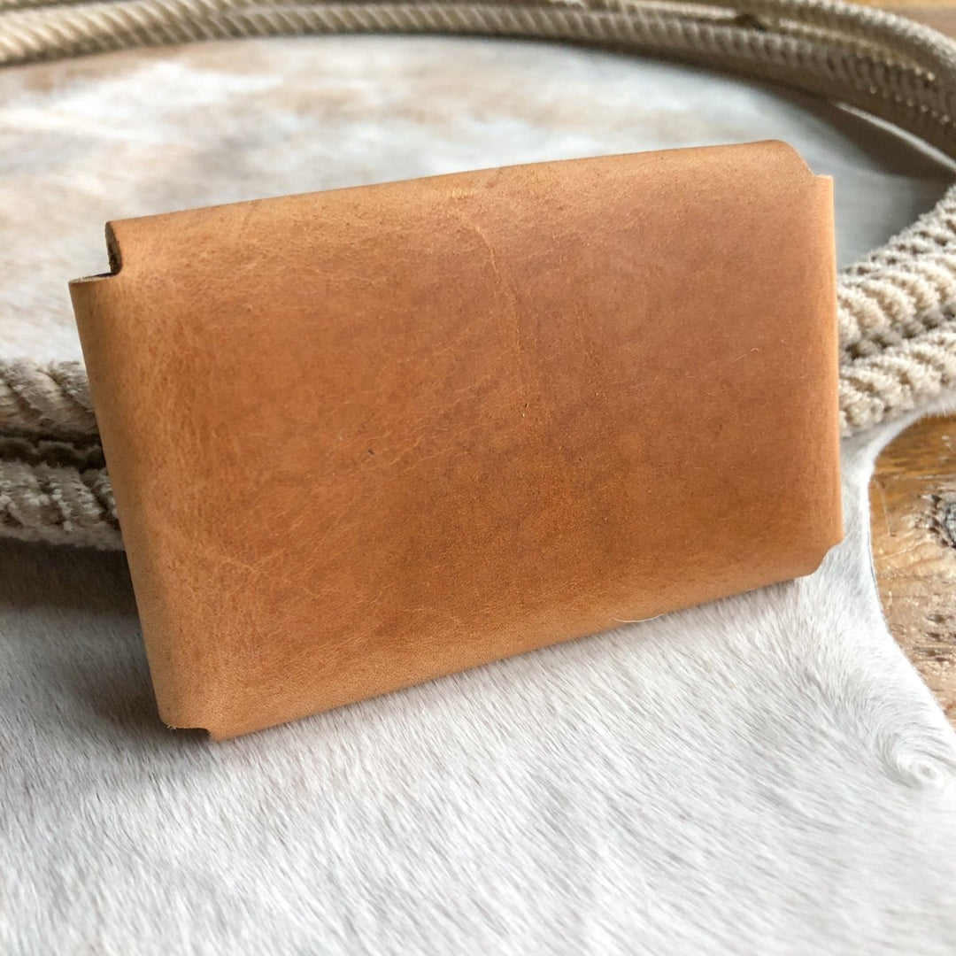 The Sorrell Card Holder by Savannah Sevens western life{&}style, in light brown Horween leather, sits on a soft white and beige textured surface with a coiled rope in the background.