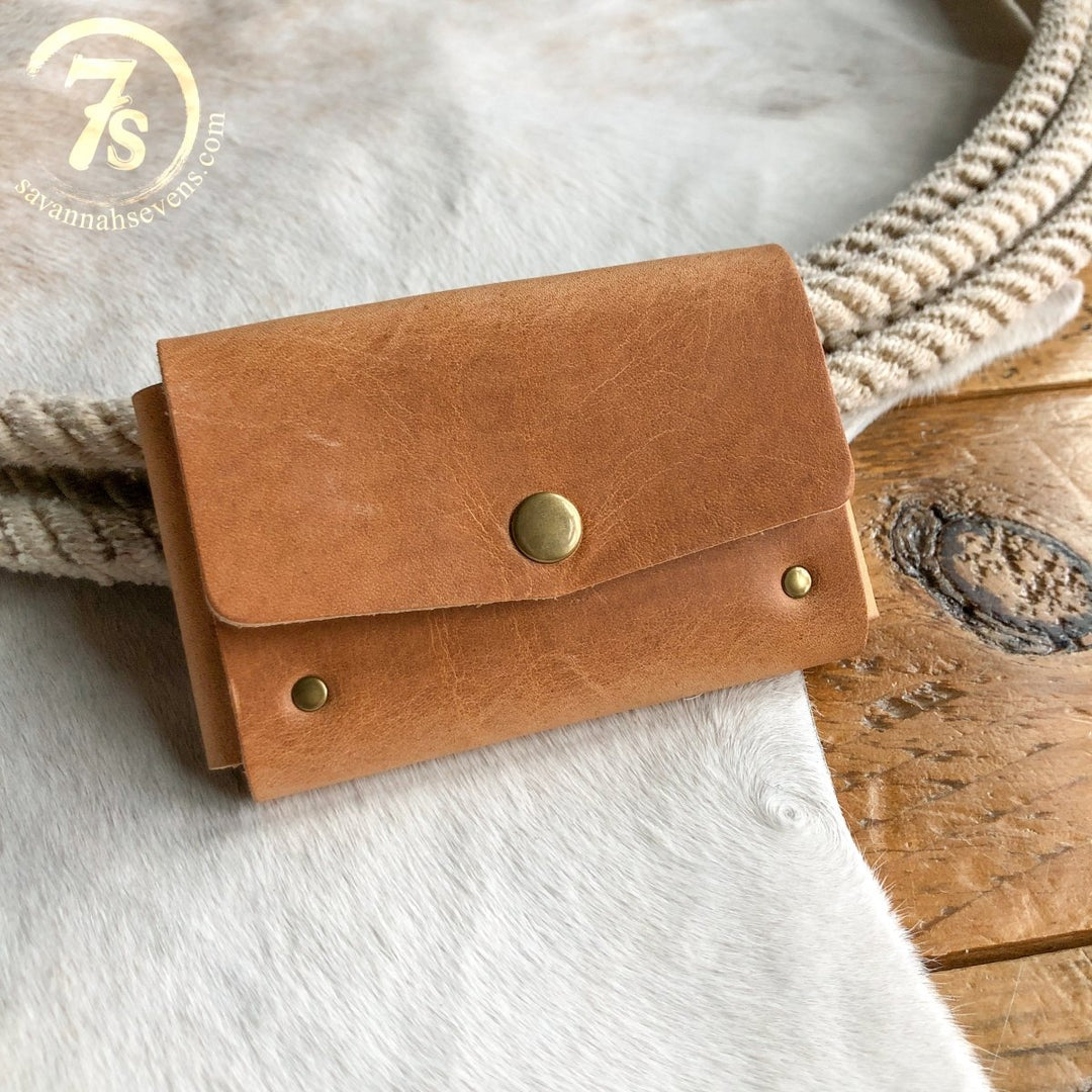 A tan Sorrell Card Holder by Savannah Sevens western life{&}style, crafted from Horween leather with a brass button and rivets, rests on a cream and tan surface beside a woven band. 7s savannahsevens.com logo appears in the top left. Made in USA.