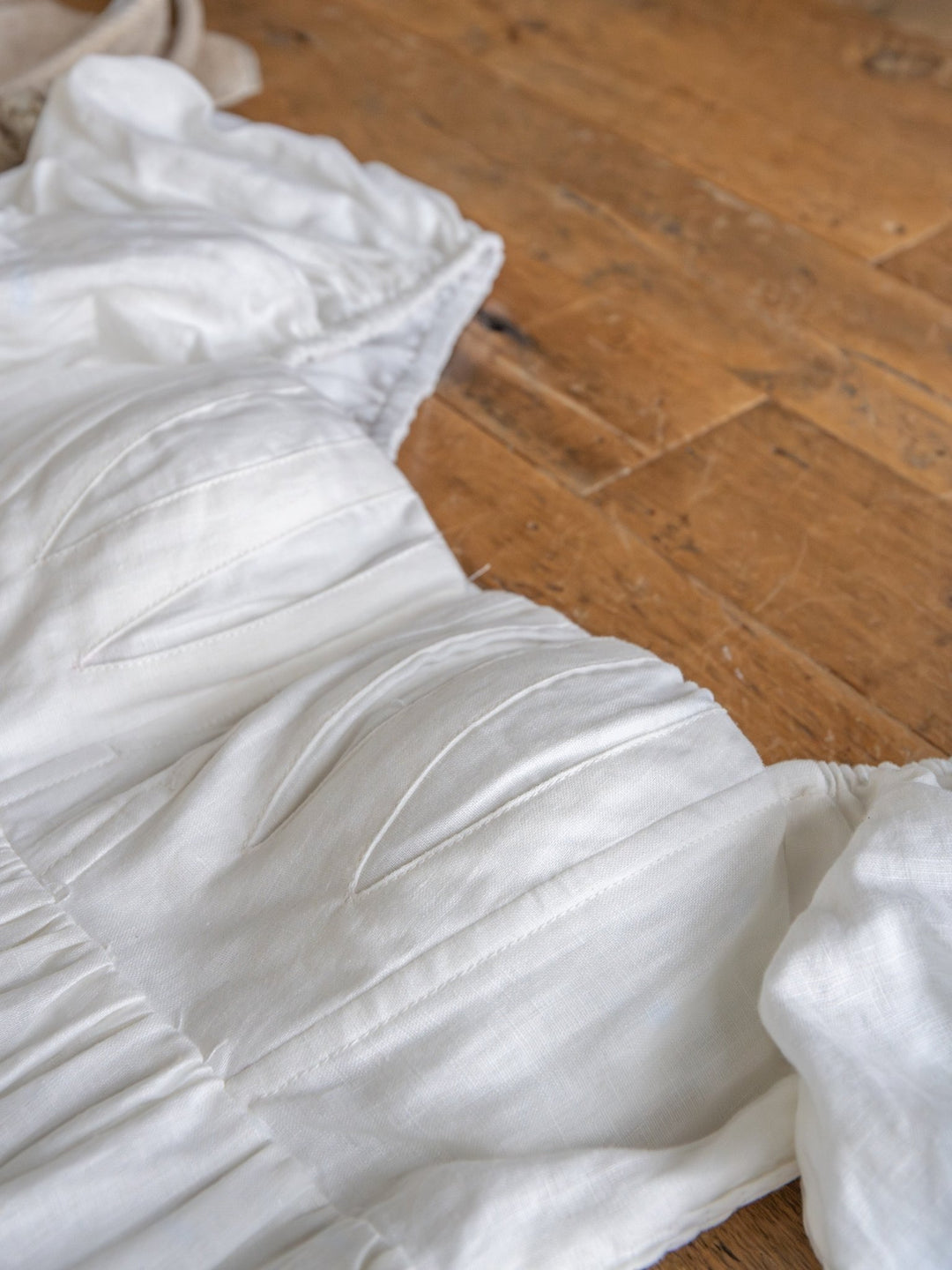 A close-up of The Southampton by Savannah Sevens western life{&}style—a white linen puff sleeve dress with ruched details, light textured fabric, and gentle folds—lying on a wooden floor.