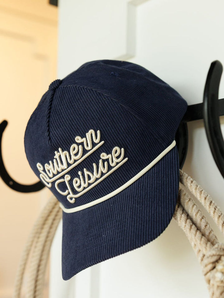 A navy corduroy Southern Leisure Cap by Savannah Sevens western life{&}style, featuring white cursive embroidery, hangs on a black hook beside a coiled rope.