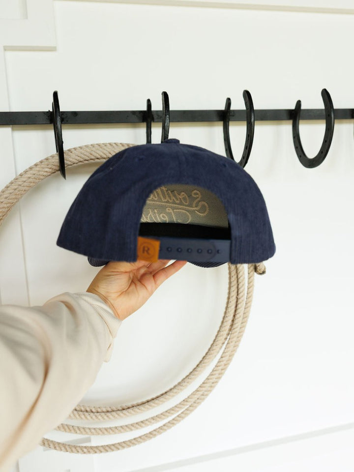 A person holds the Southern Leisure Cap by Savannah Sevens western life{&}style by its adjustable strap in front of a black wall-mounted rack with hooks and a hanging coiled rope.