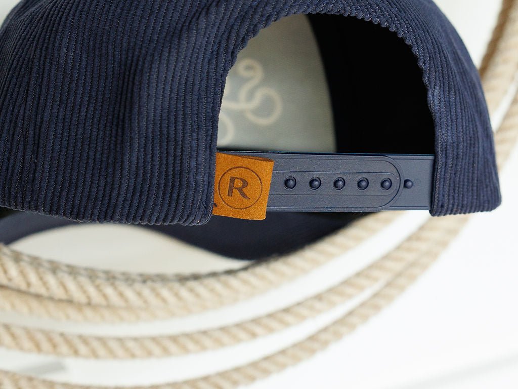 Close-up of the back of the navy Southern Leisure Cap by Savannah Sevens western life{&}style, showing its adjustable snapback and brown tag with a circled R. Thick white rope appears in the background.