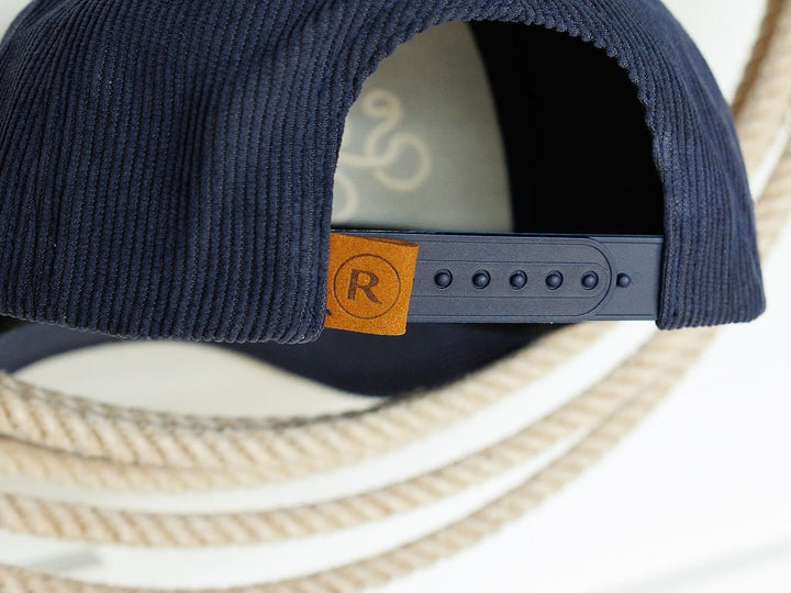 Close-up of the back of the navy Southern Leisure Cap by Savannah Sevens western life{&}style, showing its adjustable snapback and brown tag with a circled R. Thick white rope appears in the background.