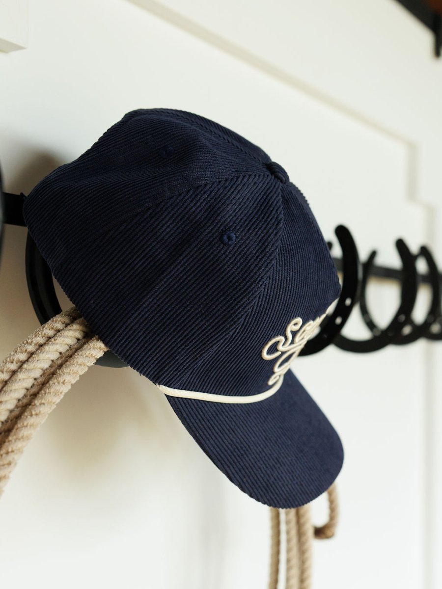 The Southern Leisure Cap by Savannah Sevens western life{&}style, a navy corduroy hat with white embroidered text, hangs on a black hook above coiled rope, with hooks mounted on a white wall in the background.