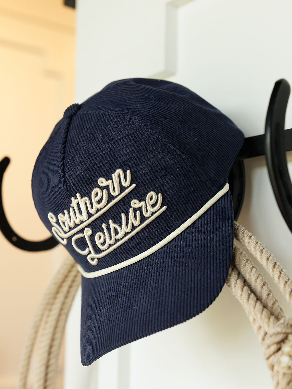 A navy corduroy Southern Leisure Cap by Savannah Sevens western life{&}style, featuring white cursive embroidery, hangs on a black hook beside a coiled rope.