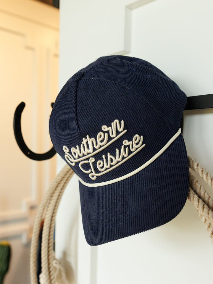 A navy Southern Leisure Cap by Savannah Sevens western life{&}style, with white embroidery, hangs on a black hook next to a coiled rope.