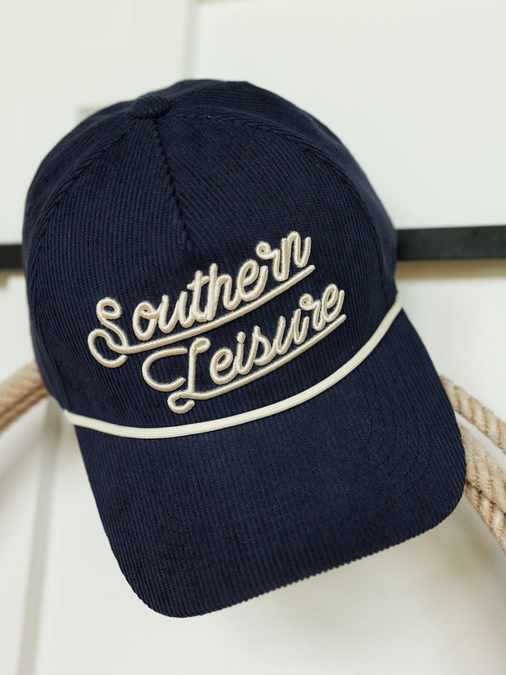 The Southern Leisure Cap by Savannah Sevens western life{&}style is a navy corduroy hat with white embroidered text, shown hanging on a hook.