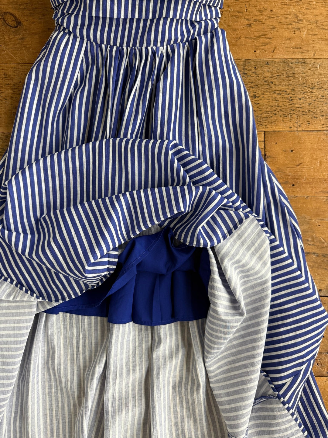 The Southport Striped Strapless Dress by Savannah Sevens western life{&}style, featuring blue and white vertical stripes, gathered fabric, and layered ruffles, is laid out on a wooden floor.