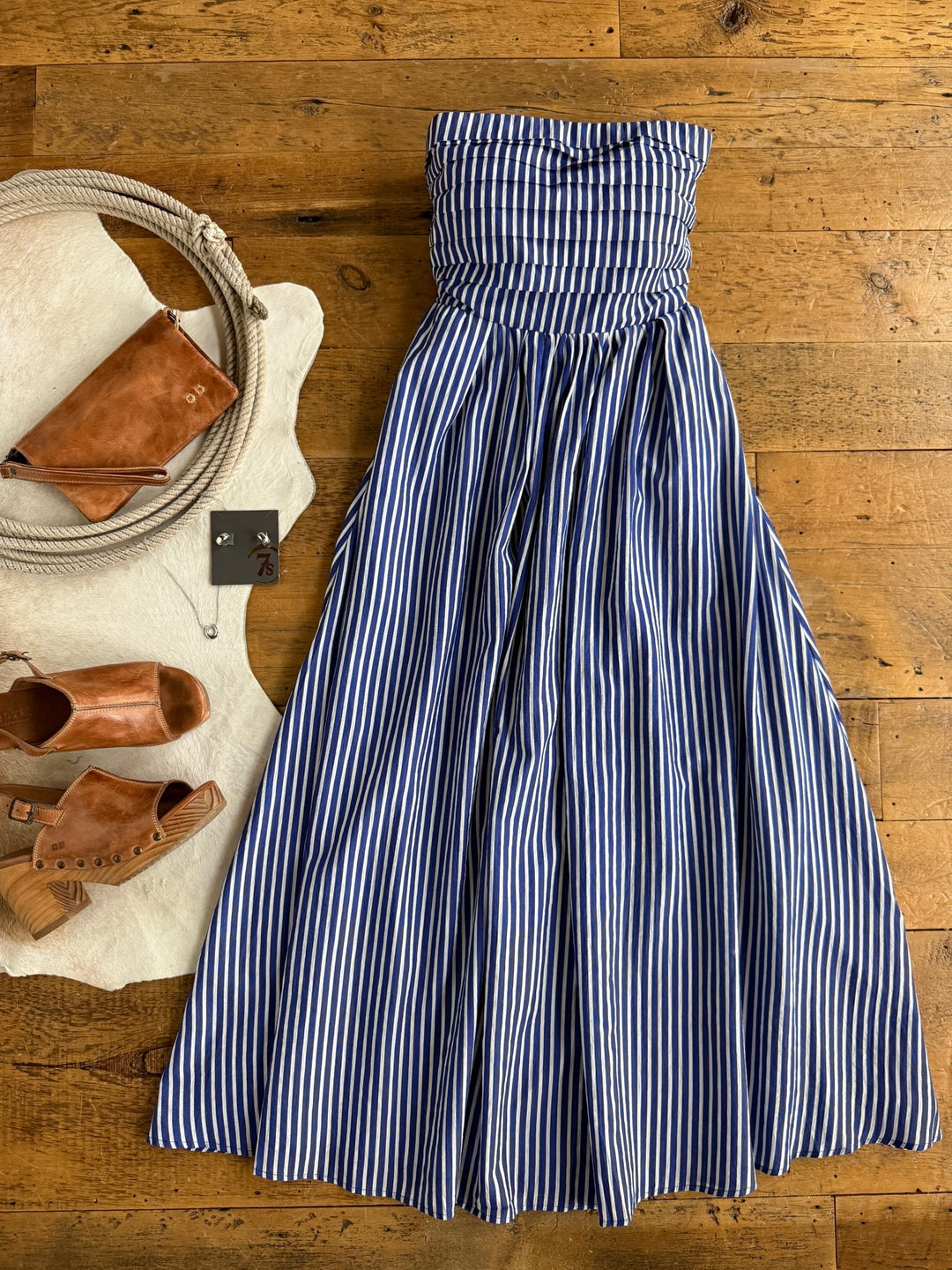 The Southport Striped Strapless Dress by Savannah Sevens western life{&}style, featuring cobalt blue and white stripes, is displayed on a wooden floor alongside brown sandals, a brown clutch, and a coiled rope basket.