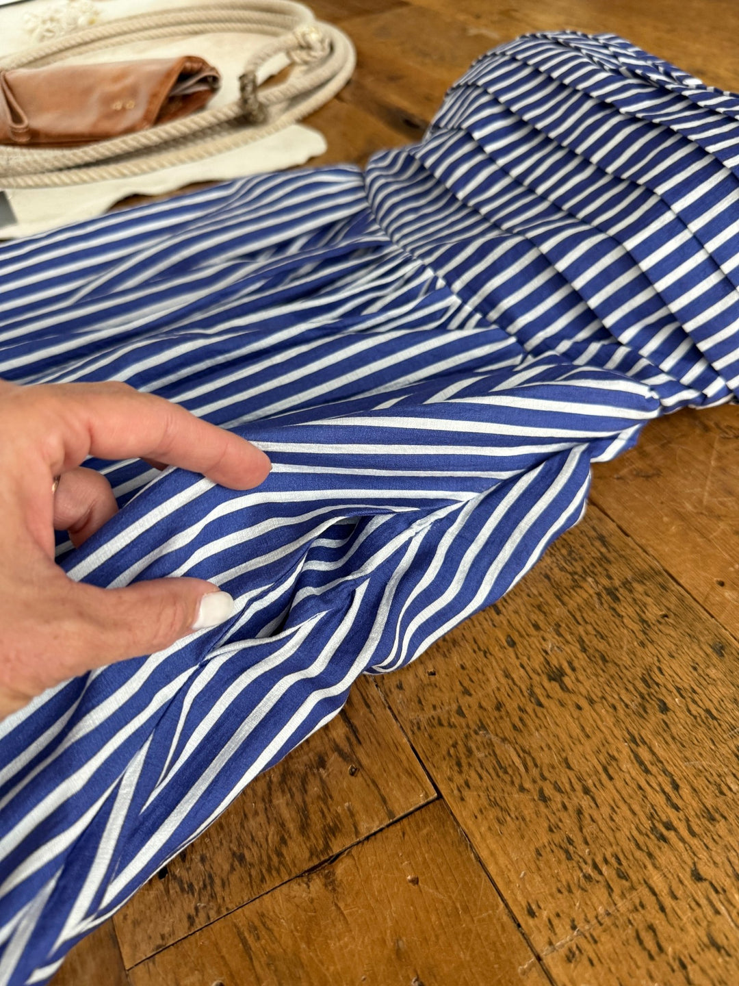 A hand with white nail polish holds the Southport Striped Strapless Dress by Savannah Sevens western life{&}style, in cobalt blue and white stripes, laid out on a wooden surface with a bag and rope accessory in the background.