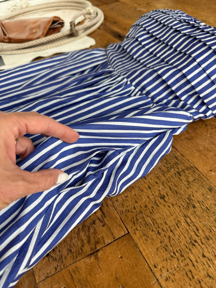 A hand with white nail polish holds the Southport Striped Strapless Dress by Savannah Sevens western life{&}style, in cobalt blue and white stripes, laid out on a wooden surface with a bag and rope accessory in the background.