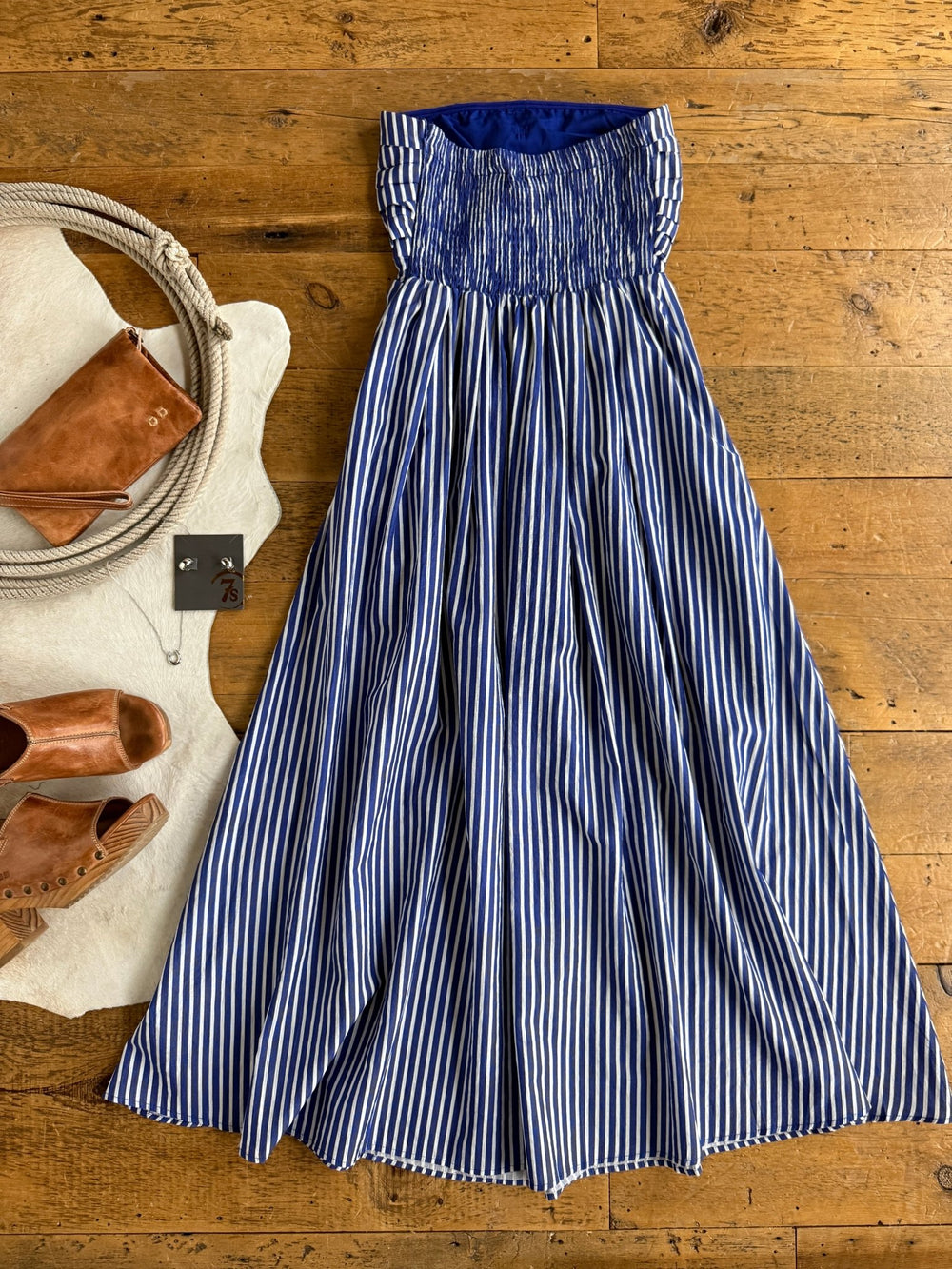 The Southport Striped Strapless Dress by Savannah Sevens western life{&}style, in cobalt blue and white stripes, is laid out on a wooden floor alongside tan sandals, a brown clutch, rope accents, and earrings.