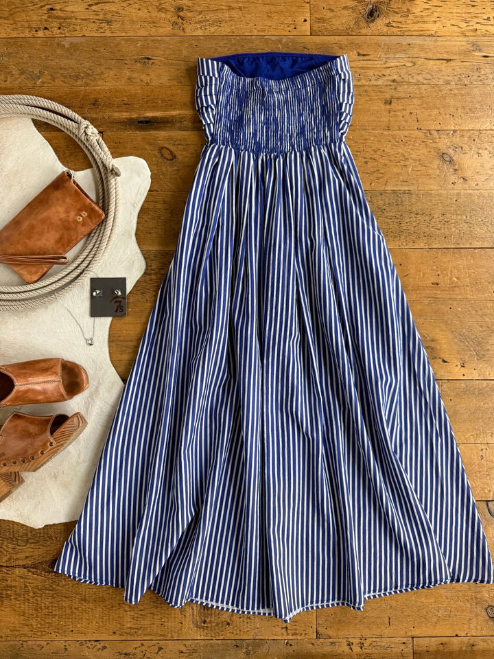 The Southport Striped Strapless Dress by Savannah Sevens western life{&}style, in cobalt blue and white stripes, is laid out on a wooden floor alongside tan sandals, a brown clutch, rope accents, and earrings.