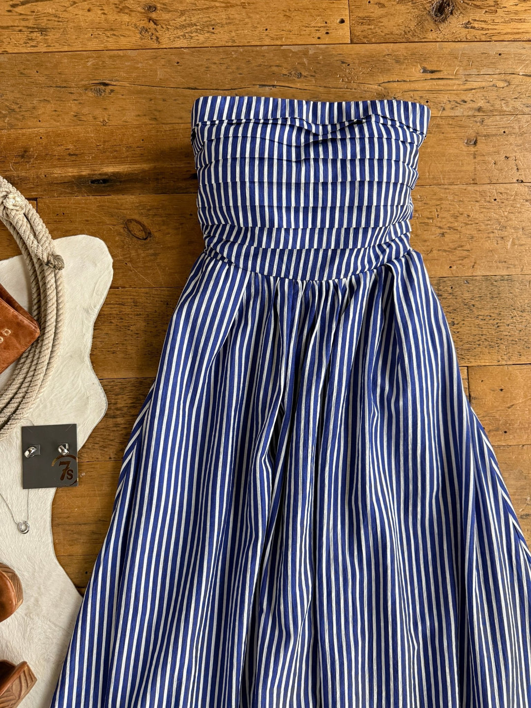 The Southport Striped Strapless Dress by Savannah Sevens western life{&}style, featuring cobalt blue and white vertical stripes, is displayed flat on a wooden floor. Nearby, various accessories such as a necklace and a leather item are partially visible.