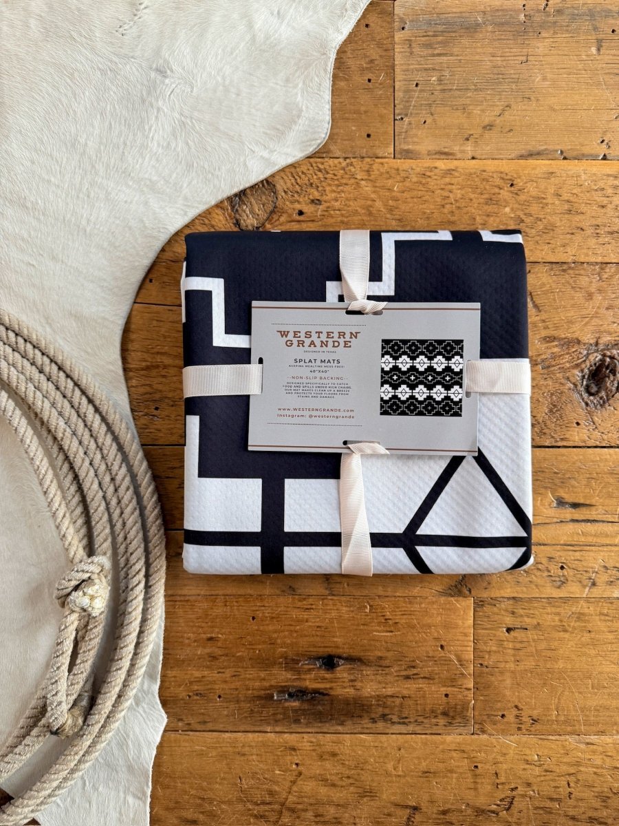 A folded black and white patterned Southwest Splat Mat by Savannah Sevens western life{&}style, made of machine washable fabric and tied with a cream ribbon, sits on a wooden floor beside a coiled rope and a white cowhide rug.