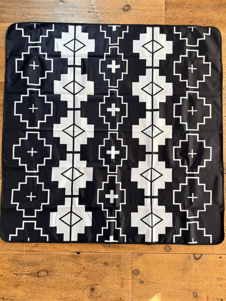 The Southwest Splat Mat from Savannah Sevens western life{&}style features a black and white symmetrical geometric pattern with squares, diamonds, and crosses. It has a nonslip backing for secure placement on wooden floors.