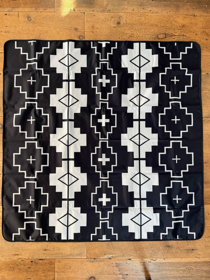 The Savannah Sevens western life{&}style Southwest Splat Mat is a square black mat with a geometric white pattern and nonslip backing, shown here on a wooden floor.