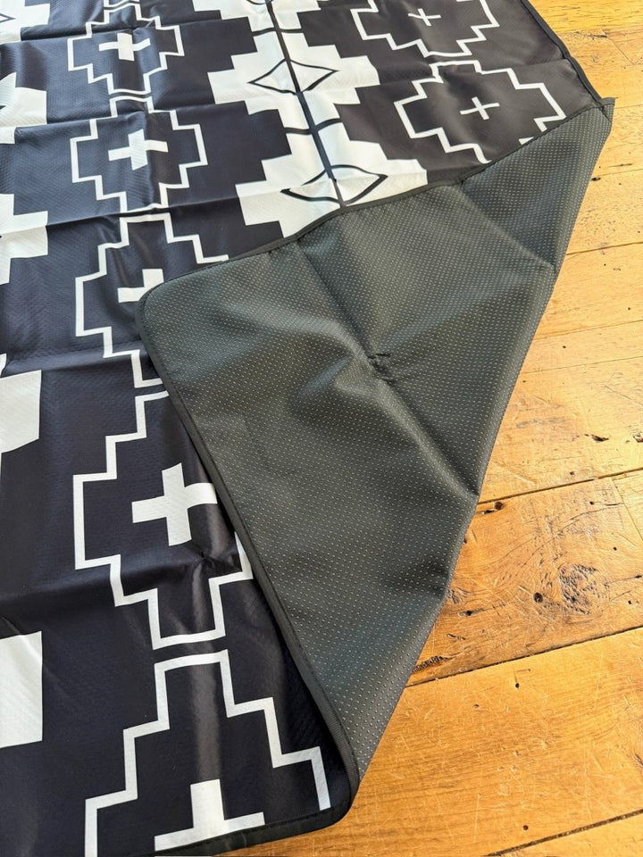 The Southwest Splat Mat by Savannah Sevens western life{&}style features a black-and-white geometric pattern, shown on a wooden floor with one corner flipped to reveal its dark, textured, nonslip backing. It's also machine washable for easy cleaning.