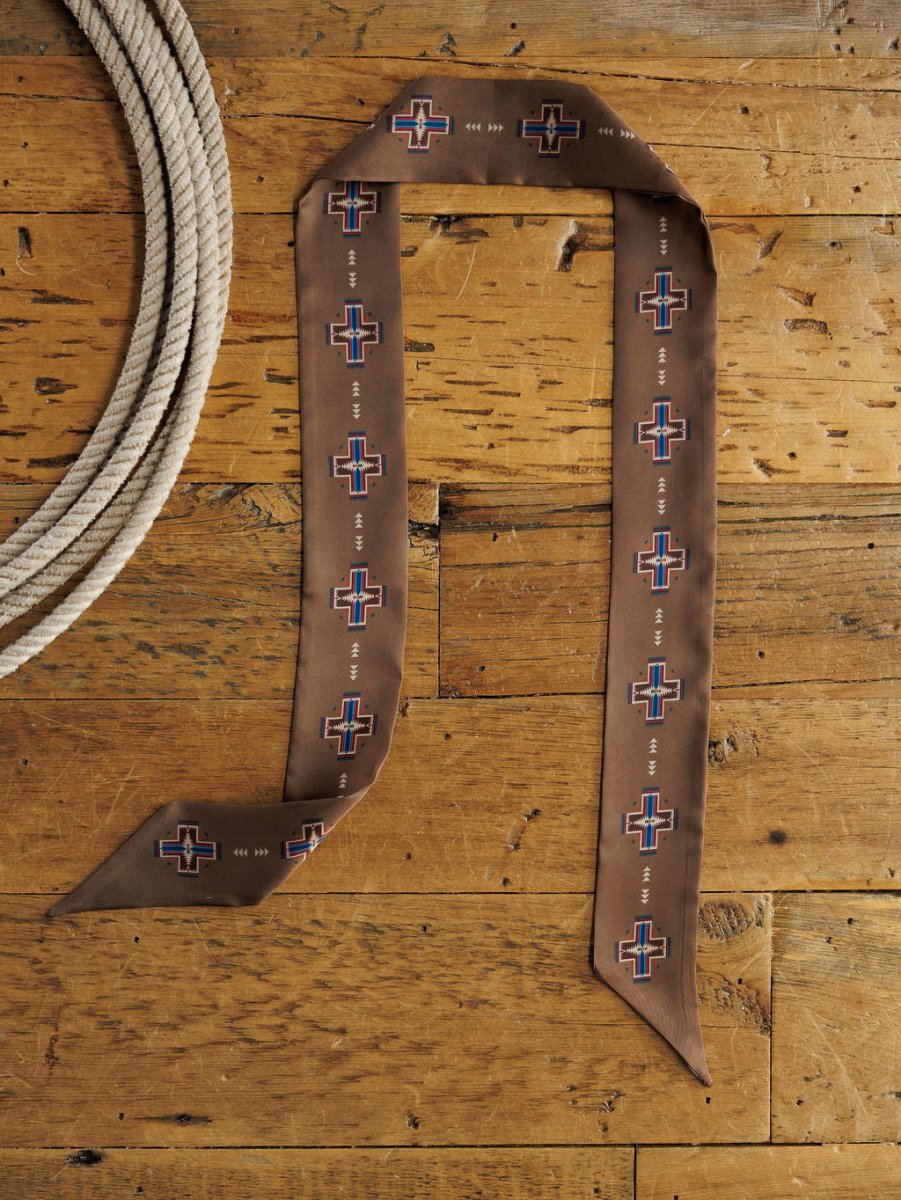 The Southwest Twilly {Brown} by Savannah Sevens western life{&}style, featuring a geometric cross pattern, is displayed in the shape of a “9” on a rustic wooden surface beside a coiled rope.