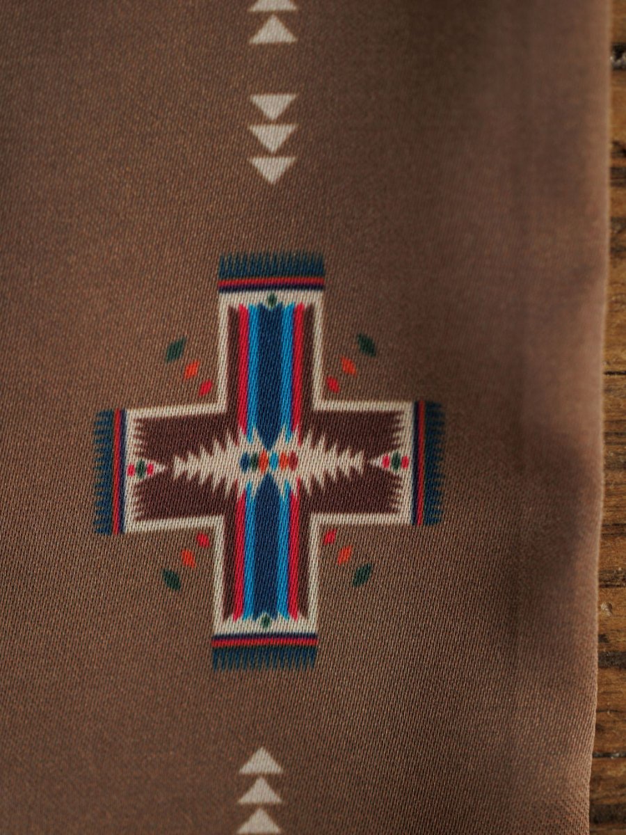 A close-up of the Southwest Twilly {Brown} scarf by Savannah Sevens western life{&}style shows a geometric cross pattern in brown, white, blue, red, and green on a brown background with part of a wooden surface at the edge.