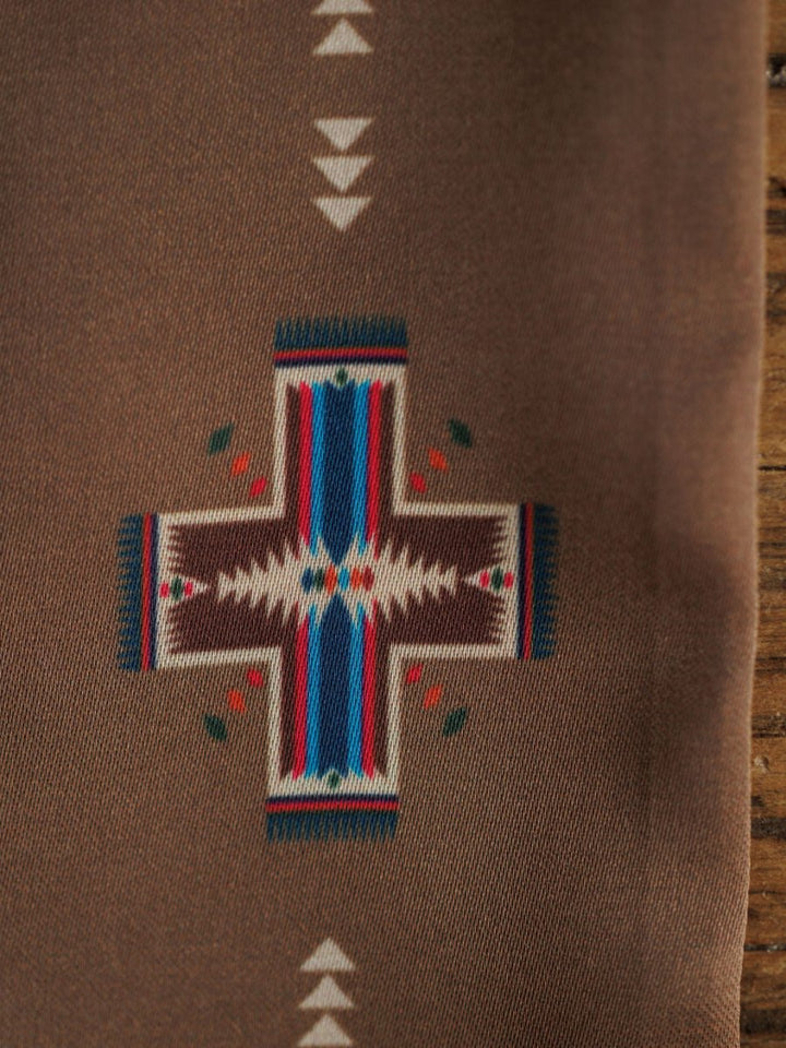 A close-up of the Southwest Twilly {Brown} scarf by Savannah Sevens western life{&}style shows a geometric cross pattern in brown, white, blue, red, and green on a brown background with part of a wooden surface at the edge.