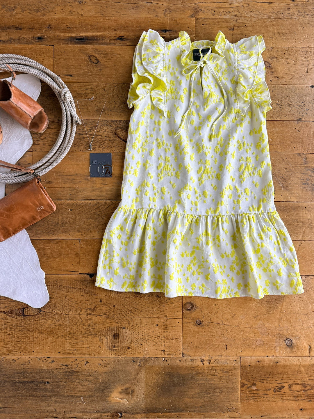 The Savannah Sevens western life{&}style Spring Valley Lemon Textured Floral Dress, featuring layered ruffle shoulders, is displayed with brown sandals, a leather purse, and a silver circular pendant necklace on a wooden floor.