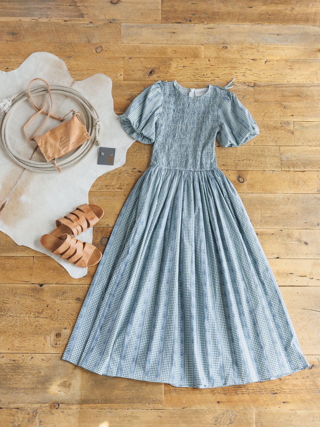 The Springfield Blue Cotton Puff Sleeve Dress by Savannah Sevens western life{&}style is laid out on a wooden floor with tan sandals, a small tan crossbody bag, and a round white rug.