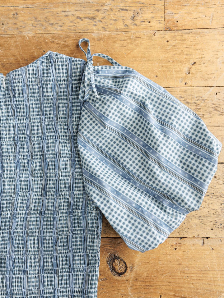 A close-up of the Springfield Blue Cotton Puff Sleeve Dress by Savannah Sevens western life{&}style, showcasing textured, smocked blue-gray fabric and a single puff sleeve with blue and white checked and striped cotton. Laid flat on wood.