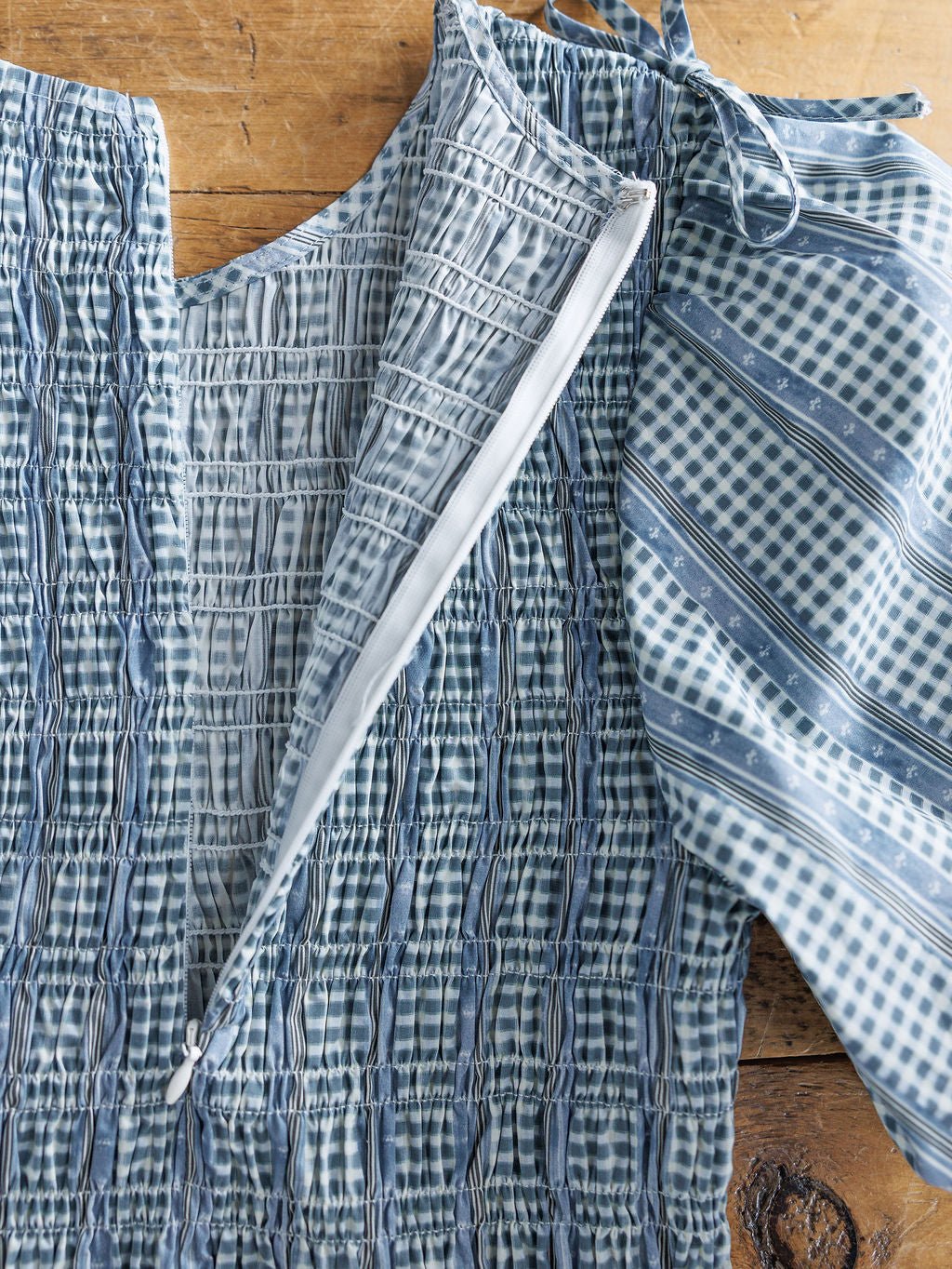 A close-up of the Springfield Blue Cotton Puff Sleeve Dress by Savannah Sevens western life{&}style, showing its blue plaid and striped cotton fabric with a visible zipper, displayed open on a wooden surface.