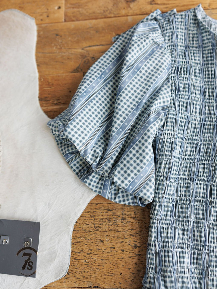 The Springfield Blue Cotton Puff Sleeve Dress by Savannah Sevens western life{&}style lies on a wooden surface beside white leather and a small grey tag labeled 7s.