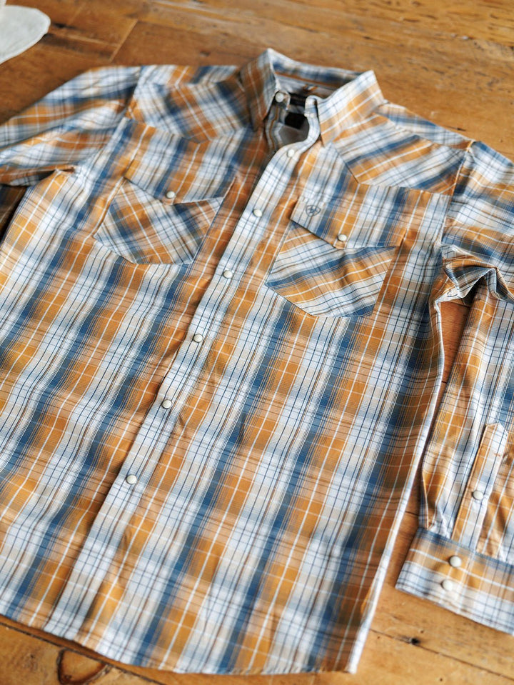A Stapleton Ariat Men's Plaid Pearl Snap shirt by Savannah Sevens western life{&}style, featuring blue, orange, and white plaid, is laid out flat on a wooden surface.