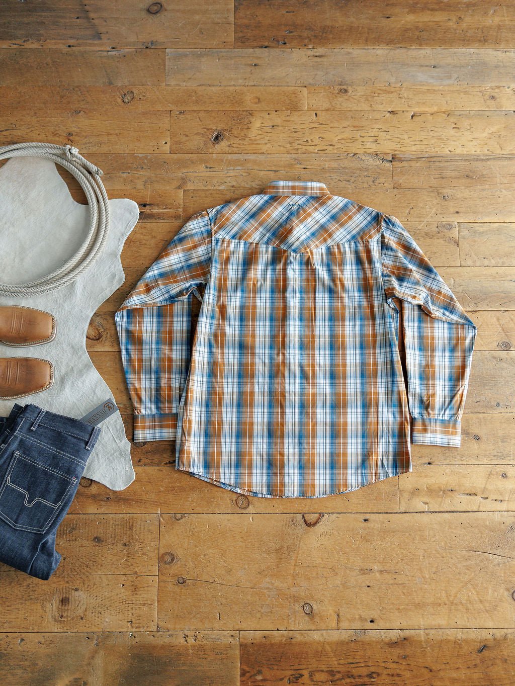 The Stapleton Ariat Men's Plaid Pearl Snap by Savannah Sevens western life{&}style, in blue, orange, and white plaid, is laid flat on a wooden floor beside blue jeans, tan shoes, a coiled rope, and a light leather hide.