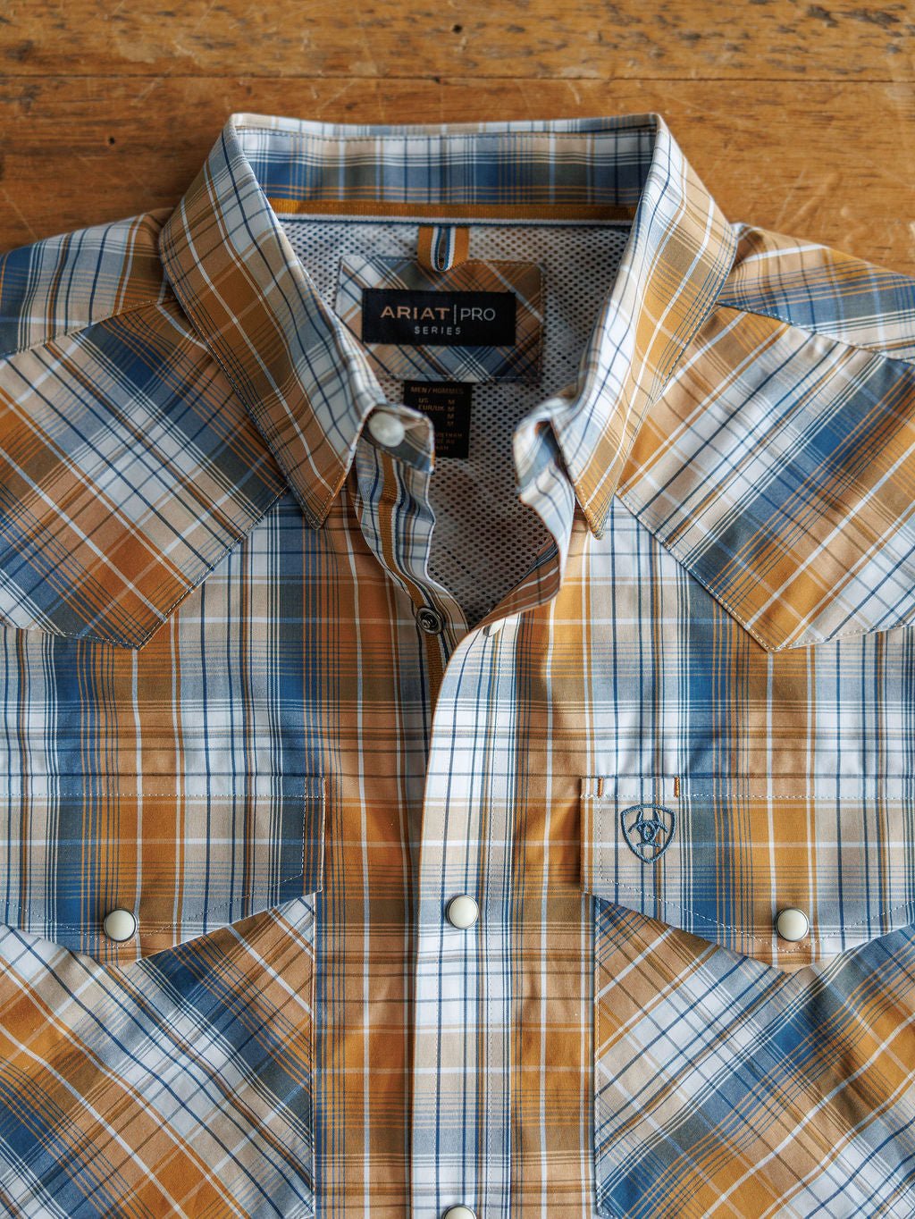 A Stapleton Ariat Men's Plaid Pearl Snap shirt by Savannah Sevens western life{&}style, featuring blue, orange, and white plaid, is folded and buttoned up on a wooden surface.