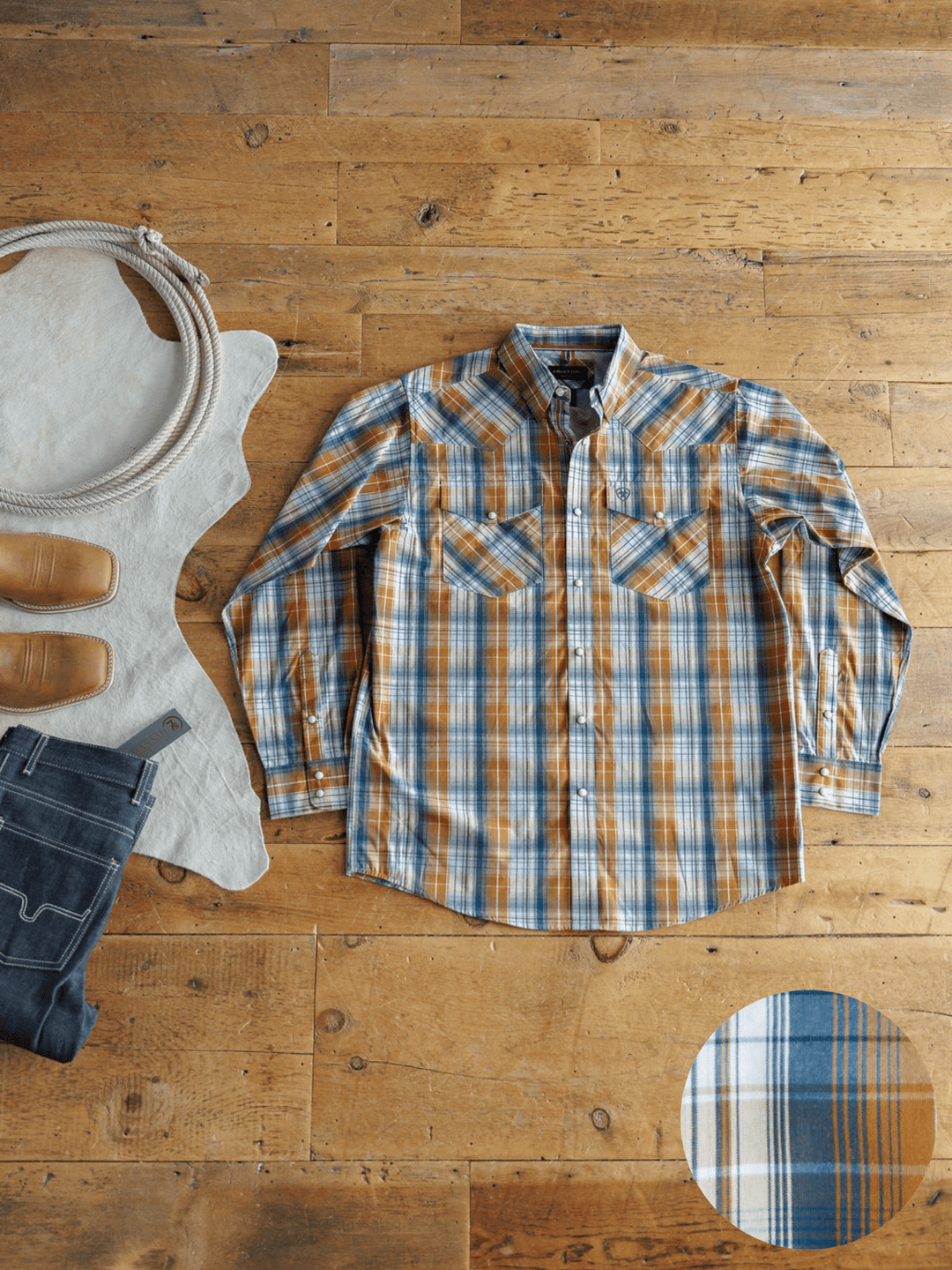 The Stapleton Ariat Men's Plaid Pearl Snap by Savannah Sevens western life{&}style is laid flat with brown boots, blue jeans, a coiled rope, and a light cowhide. A close-up shows the shirt’s blue, orange, and white plaid pattern.