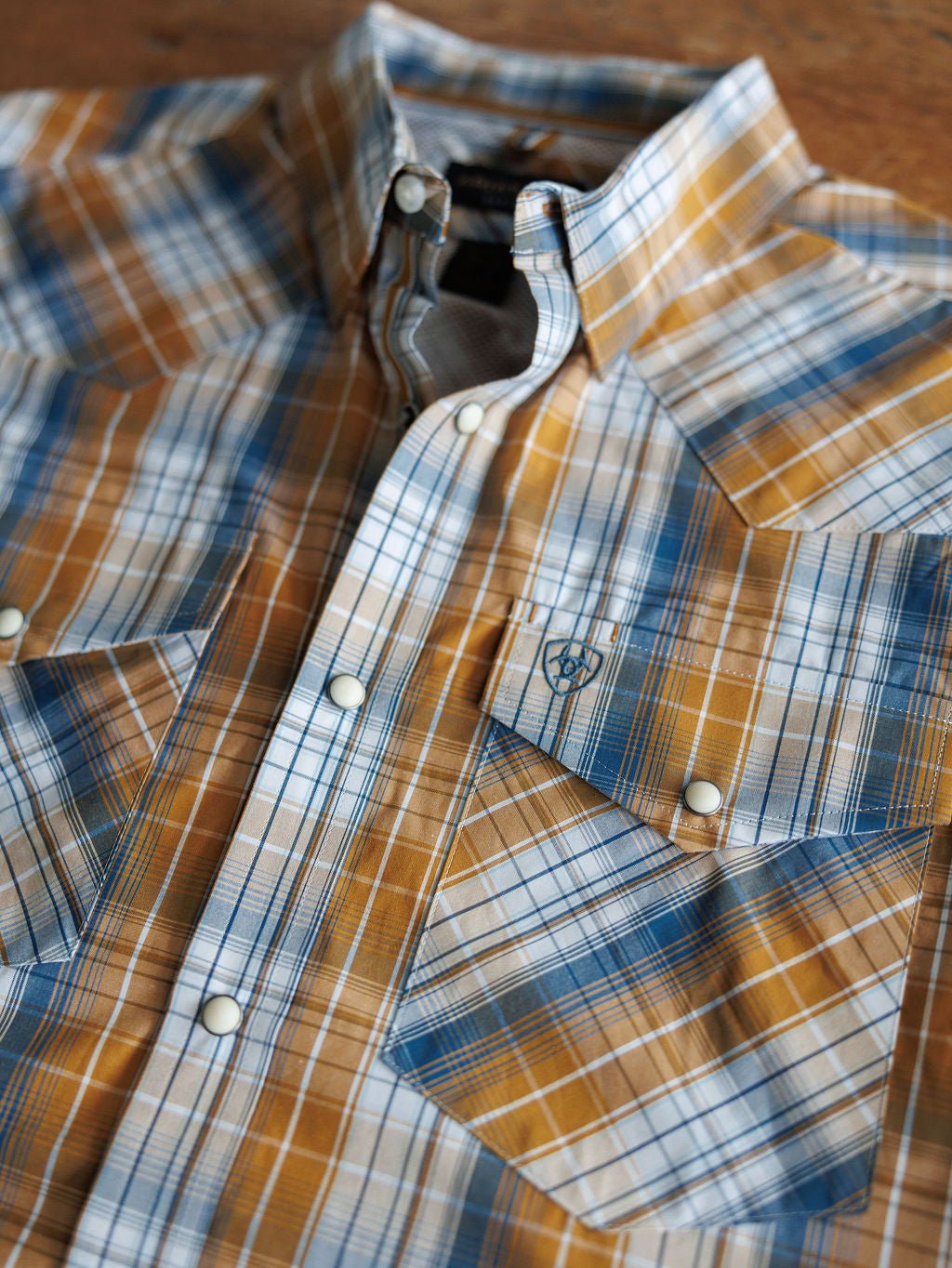 A close-up of the Stapleton Ariat Men's Plaid Pearl Snap by Savannah Sevens western life{&}style in blue, orange, and white plaid with pearl snaps and pointed flap pockets, laid flat on a wooden surface.