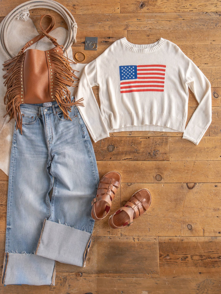 Flat lay: light-wash cuffed jeans, The Stars & Stripes long sleeve by Savannah Sevens western life{&}style, brown fringe bag, tan sandals, gold bracelet, and beige hat on a wooden floor.