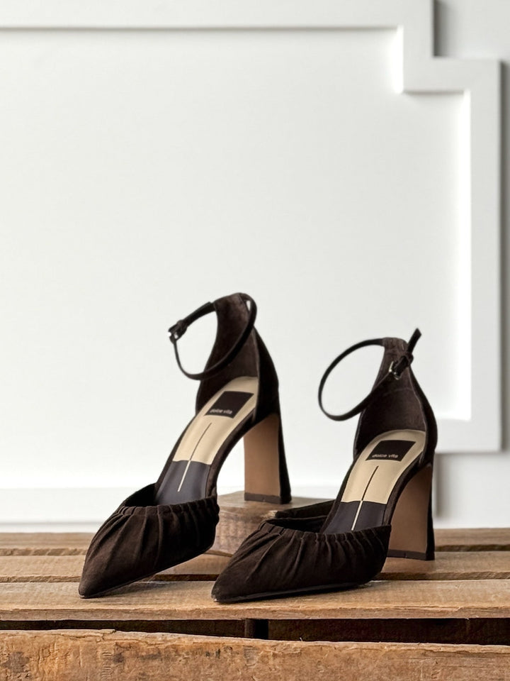 The Stella Espresso Suede Heel by Savannah Sevens western life{&}style is a pair of black high-heeled shoes featuring adjustable buckle ankle straps and ruched pointed toes, elegantly displayed on a rustic wooden surface against a white textured wall.