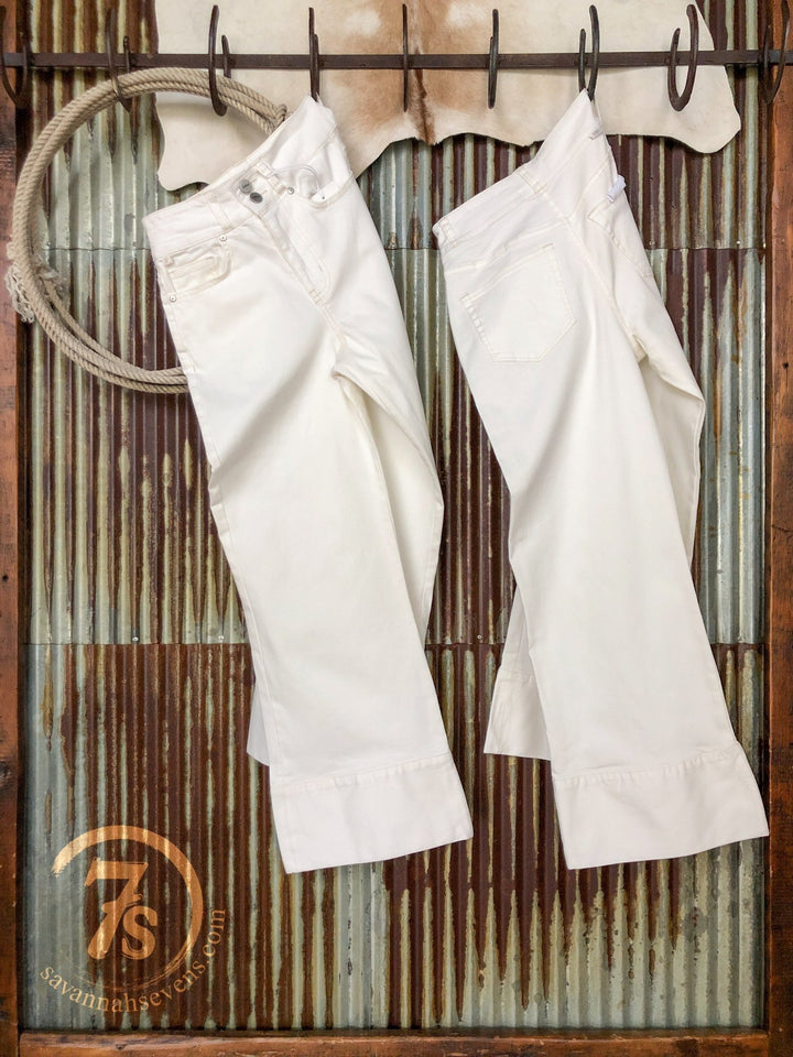 Two pairs of The Sterling High Rise Wide Leg Crop Jeans by Savannah Sevens western life{&}style hang on hooks before a rusted corrugated metal wall with a cowhide above. A circular 7s logo appears in the bottom left corner.