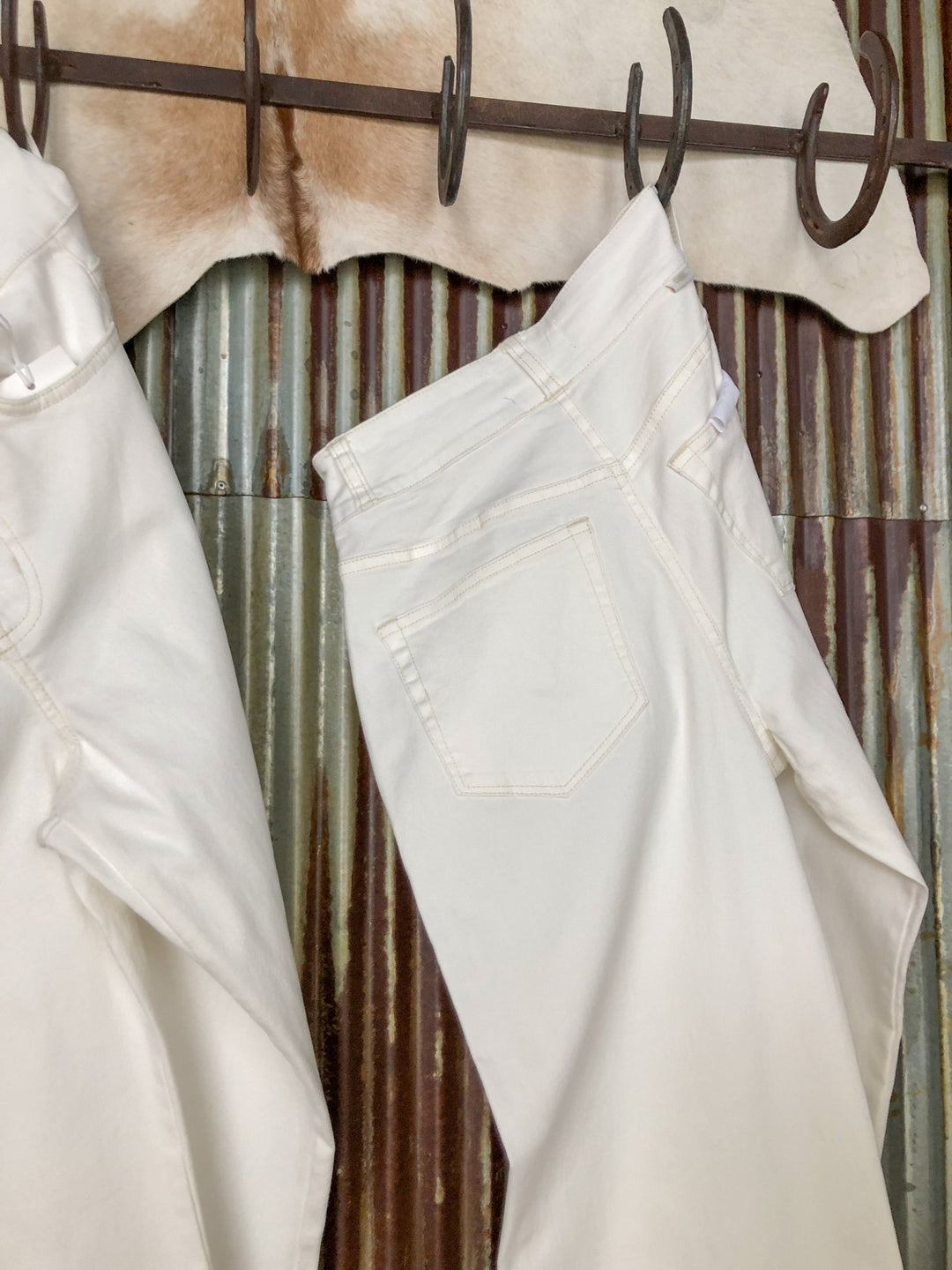 Two pairs of white denim jeans by Savannah Sevens western life{&}style hang on a metal hook rack with cowhide above. One is The Sterling High Rise Wide Leg Crop Jean, while the other features a wide leg silhouette.