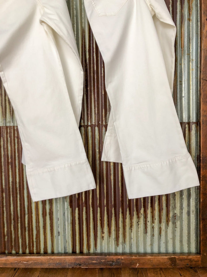 Two pairs of The Sterling High Rise Wide Leg Crop Jean by Savannah Sevens western life{&}style hang side by side, showing only the lower legs and hems in front of a rustic, vertically ridged metal and wood wall.