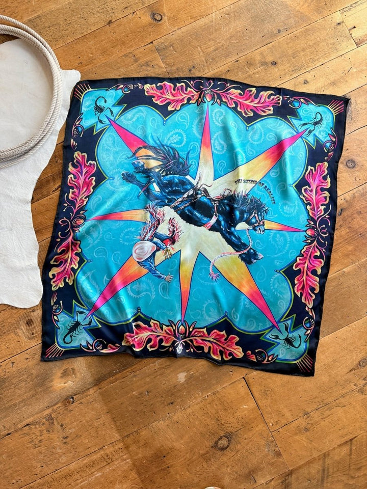 The Sting Of Reality Wild Rag by Savannah Sevens western life{&}style, featuring a bold blue base with red and yellow starburst and cowboy details, is displayed on a wooden floor beside a white rug.