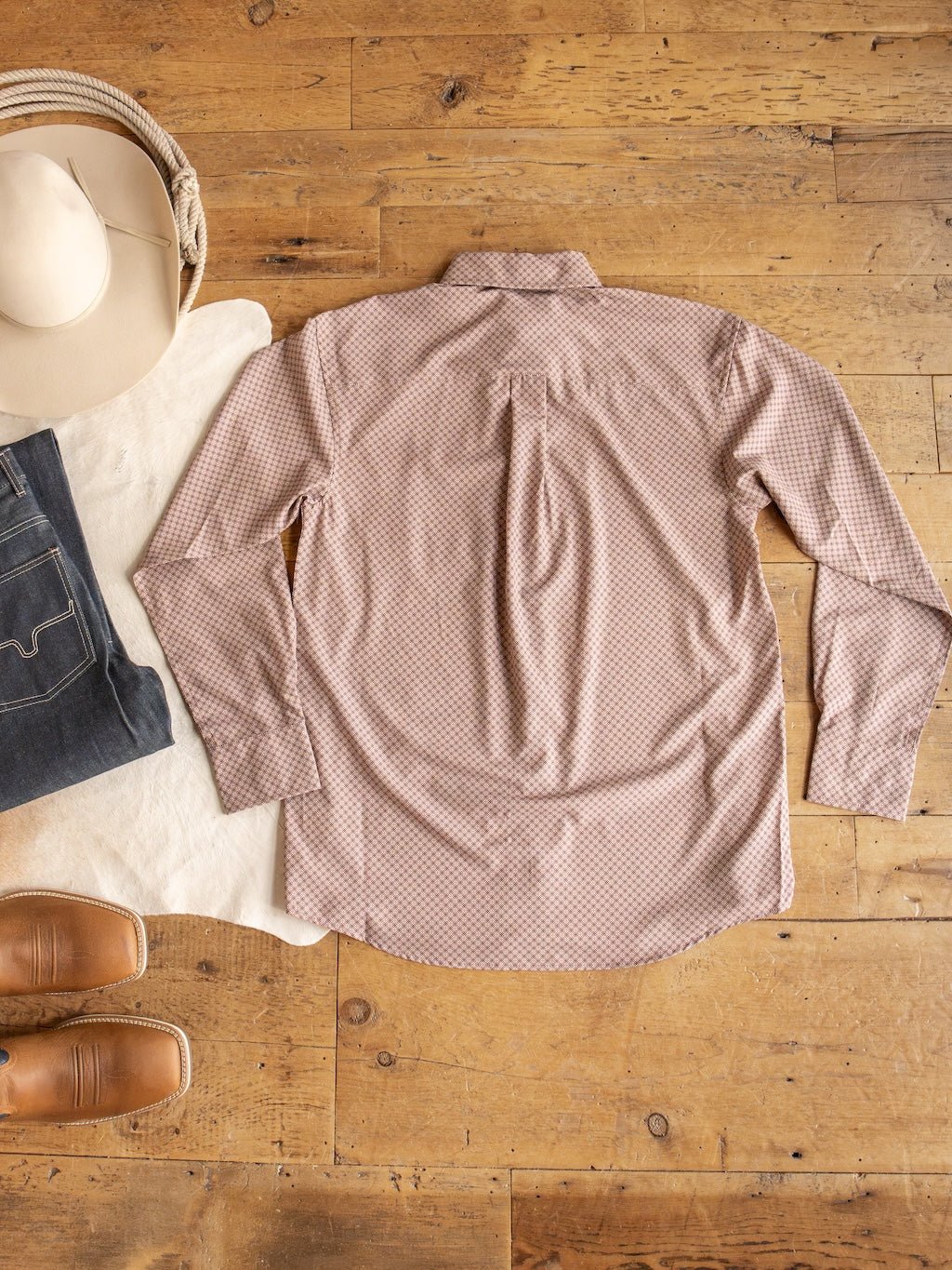 A pink patterned Strait Wrangler Men's Performance Button Up from Savannah Sevens western life{&}style is laid flat on a wooden floor, displayed alongside jeans, brown boots, a cowboy hat, and a cream-colored cloth.
