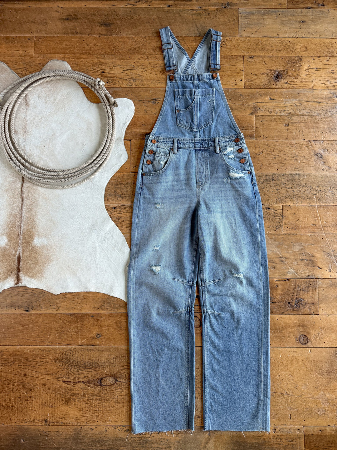 The Stran Denim Barrel Overalls by Savannah Sevens western life{&}style, in a light blue distressed wash with adjustable straps, are laid flat on a wooden floor beside a coiled rope atop a white cowhide rug.
