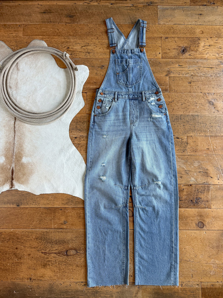 The Stran Denim Barrel Overalls by Savannah Sevens western life{&}style, in a light blue distressed wash with adjustable straps, are laid flat on a wooden floor beside a coiled rope atop a white cowhide rug.