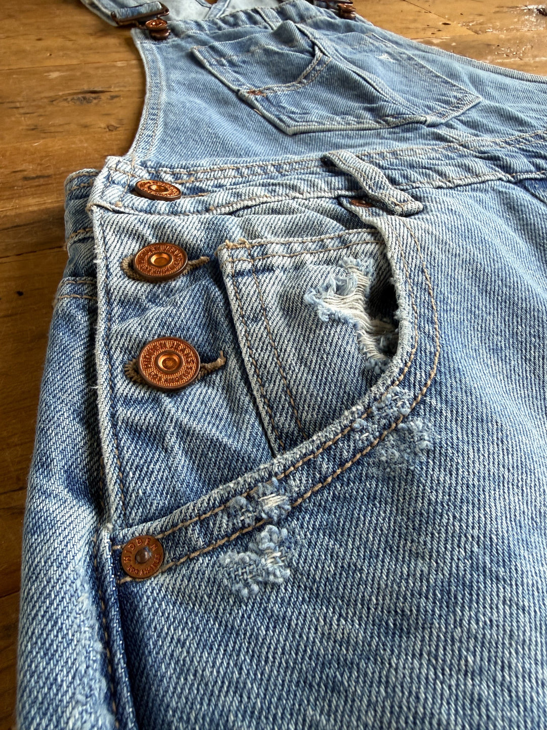 Close-up of the Stran Denim Barrel Overalls by Savannah Sevens western life{&}style, featuring distressed denim with copper buttons and adjustable straps, laid flat on a wooden surface. Visible torn sections and worn fabric highlight their vintage-inspired, well-worn appearance.