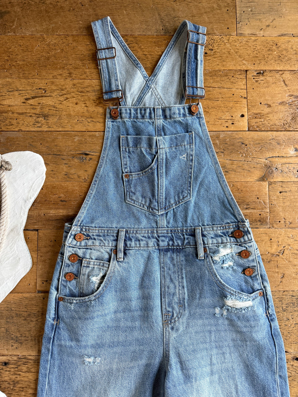 The Stran Denim Barrel Overalls by Savannah Sevens western life{&}style, featuring adjustable straps, a front chest pocket, side button details, and subtle distressing, are displayed flat on a wooden surface.