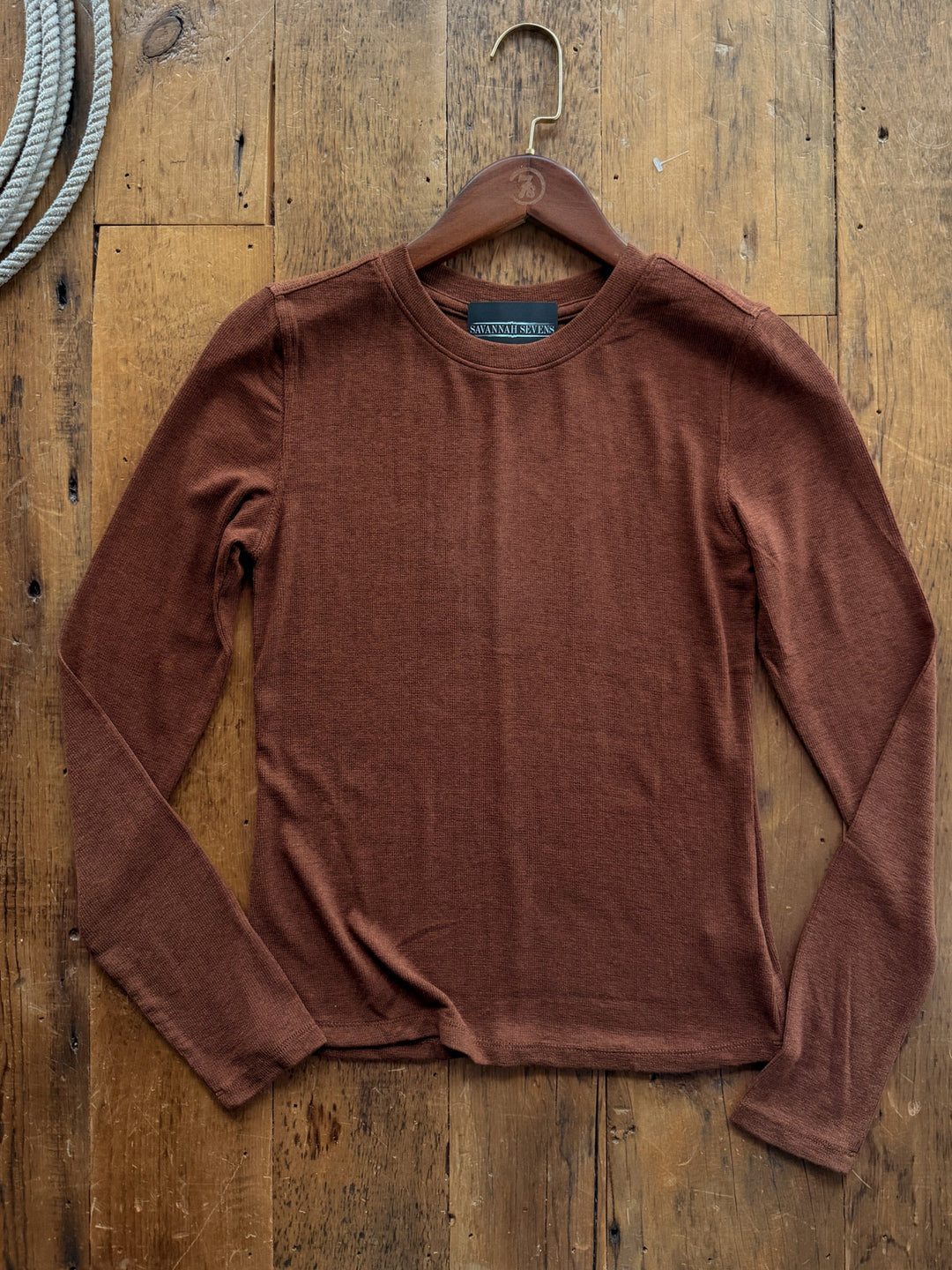 A chocolate ribbed Stratford Chocolate Ribbed Basic Top with long sleeves and a crew neck on a wooden hanger against a wooden background.