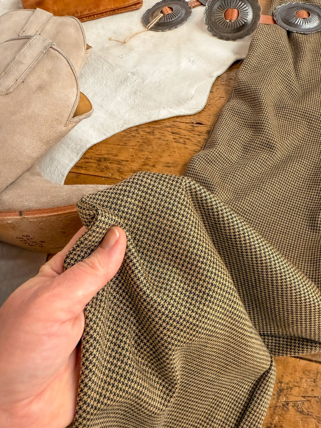 A hand displays brown and black houndstooth fabric beside tan suede boots and metallic conchos, capturing the timeless style of the Stratford Houndstooth Jumpsuit by Savannah Sevens western life{&}style.
