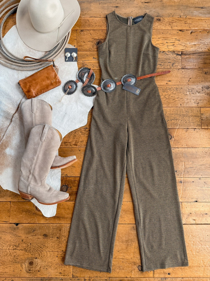Flat lay of the Savannah Sevens western life{&}style Stratford Houndstooth Jumpsuit with a concho belt, beige cowboy boots, tan cowgirl hat, brown clutch, and beaded earrings on a wooden floor with a cowhide accent.